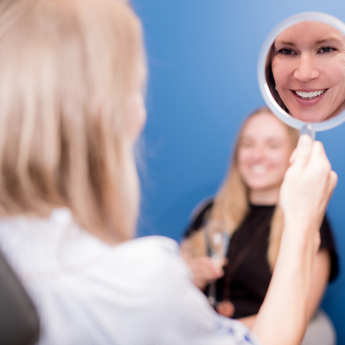 Frau schaut in einen Spiegel während im Hintergrund Freundin ein Glas Sekt trinkt