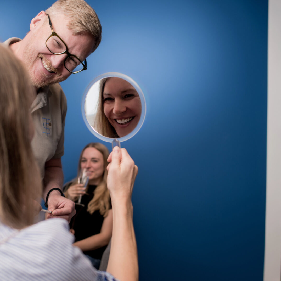 Frau schaut in einen Spiegel während im Hintergrund Freundin ein Glas Sekt trinkt und Dr. Schmidt sie anlächelt